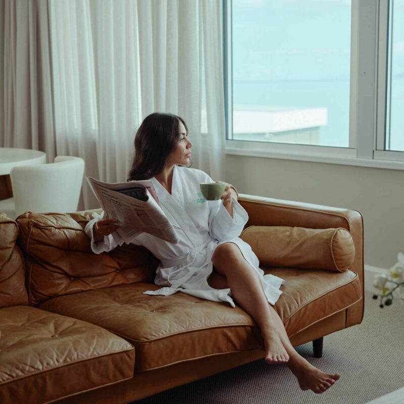 Woman in a hotel room sitting on a leather sofa, holding a coffee and reading a newspaper while looking out to ocean views.