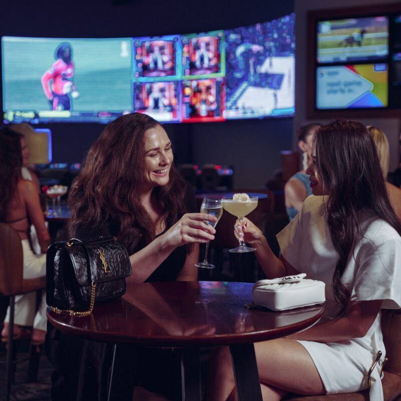 Two women enjoying cocktails at Sports Bar inside the casino, with screens and a lively atmosphere