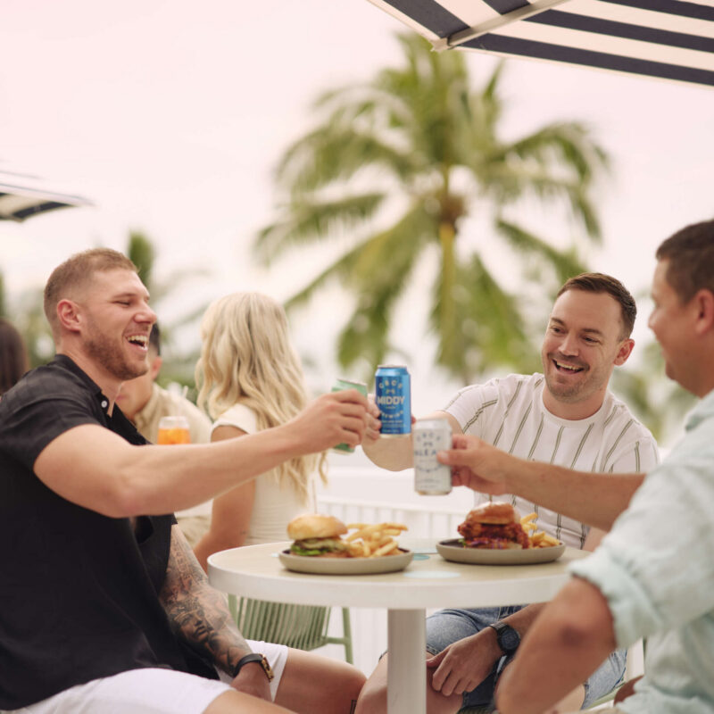 Group of men enjoying drinks and food at an outdoor table at Quarterdeck, with palm trees and waterfront views.