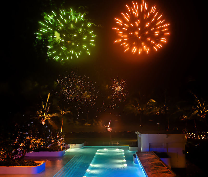 Fireworks lighting up the night sky over a pool and tropical waterfront setting